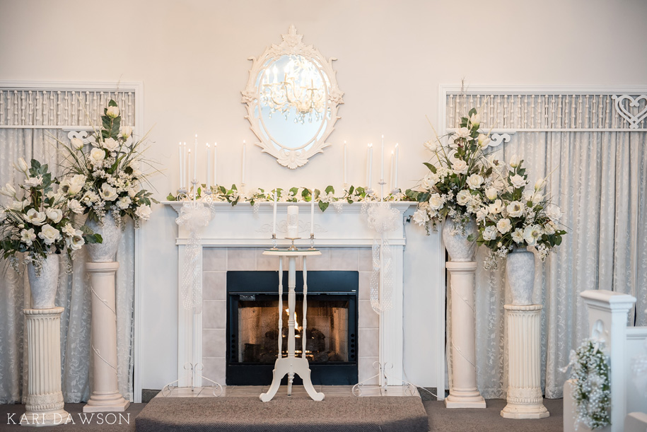 Wedding Ceremony Arrangements: The altar is flanked by white urns that hold classic arrangements of white florals. Mantle is adorned with a drape of florals in white. Finishing touches include an elegant mirror, crystal chandelier and baby's breath wreaths.