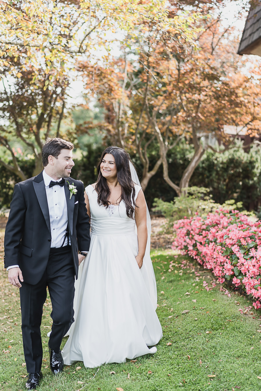 The couple smiles at each other while strolling through colorful autumn trees and pink flowers during their black-tie Michigan wedding.