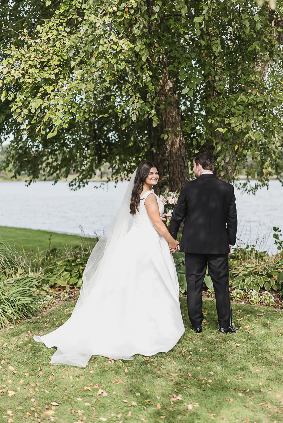 The bride smiles sweetly toward the camera while holding hands with her groom beneath tall trees along the lakefront.