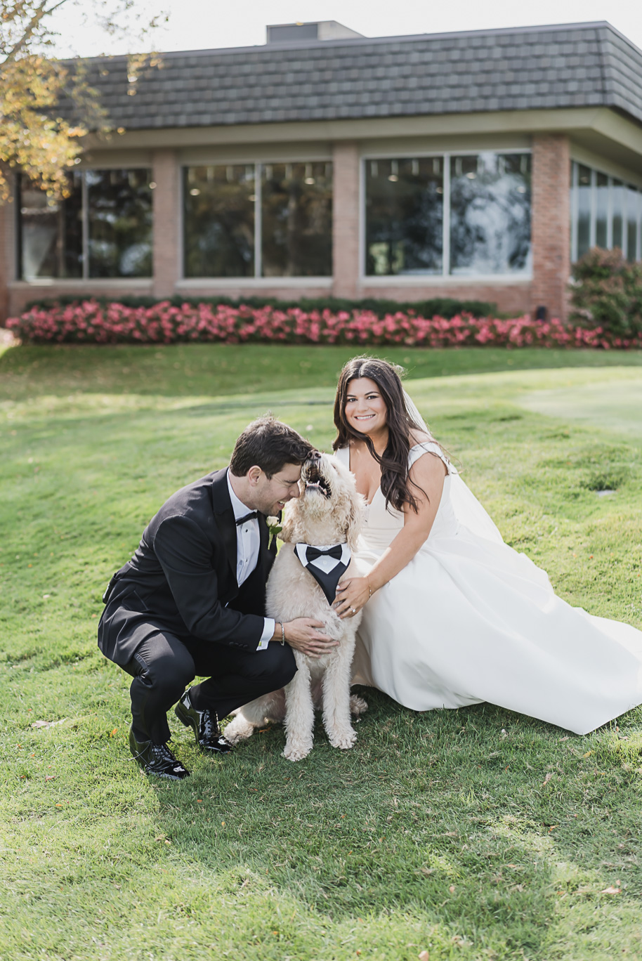The couple walks hand in hand with their doodle wearing a bow tie, adding personality and charm to their Bay Pointe Golf Club wedding portraits.