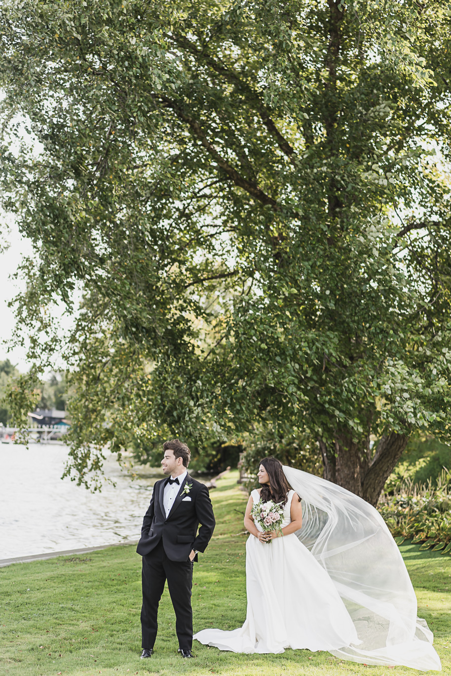 The bride and groom pose by the lake as her cathedral veil blows in the wind, creating a dramatic black-tie wedding portrait.