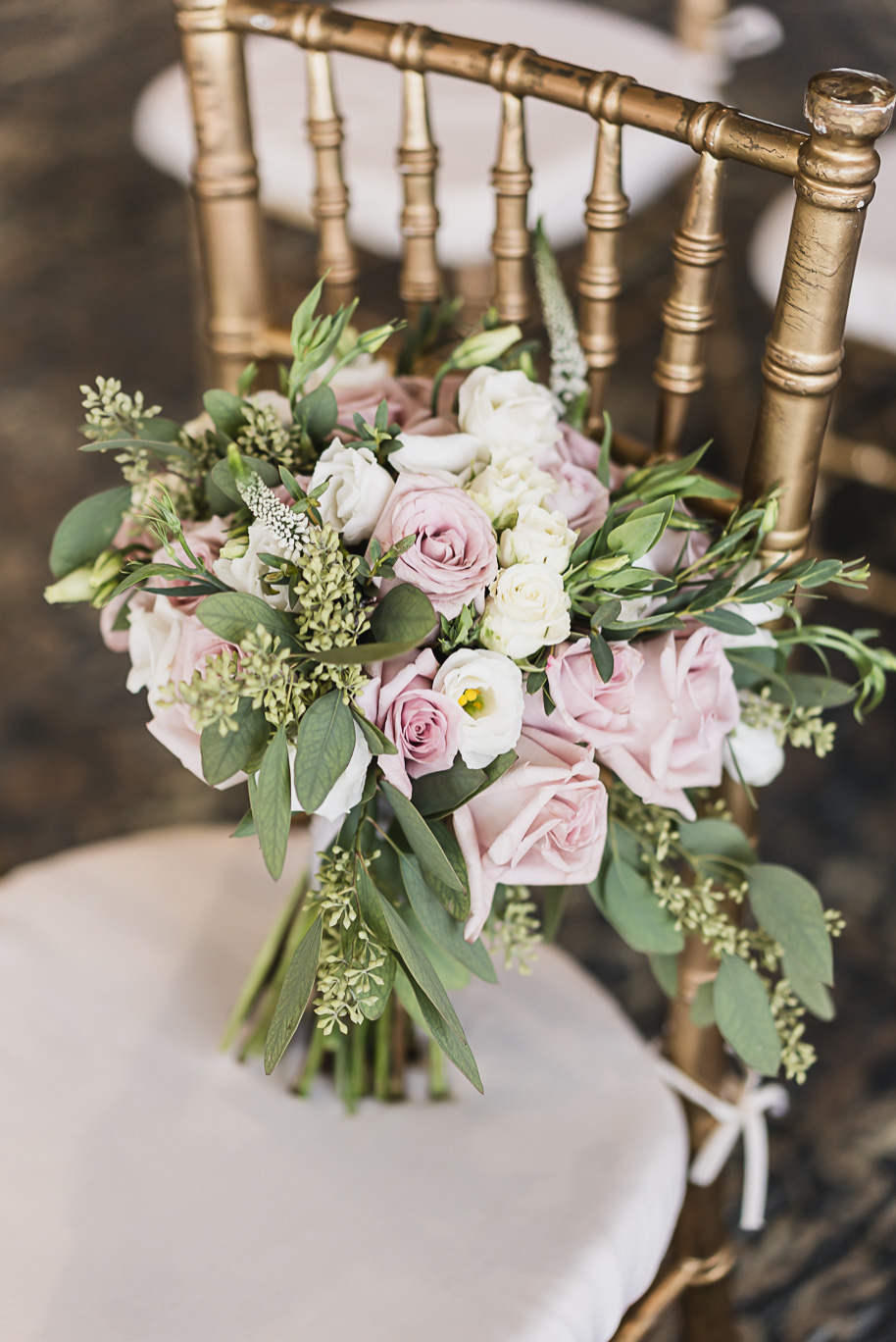 Lush bridal bouquet of blush and white roses with greenery placed on a gold chiavari chair at Bay Pointe wedding reception.