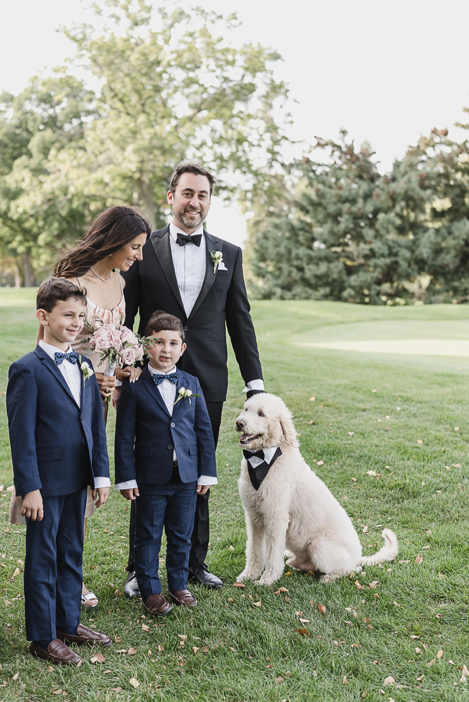 A joyful family photo including two ring bearers, a groomsman, and the couple’s dog in a bow tie, captured on the golf course lawn.