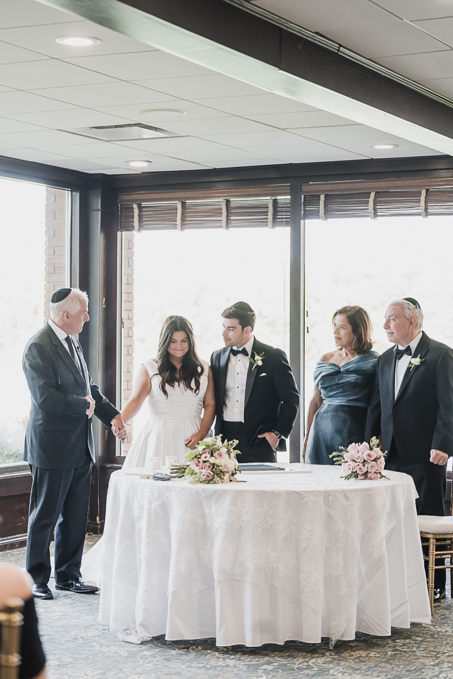 Bride and groom stand hand in hand during their Jewish ketubah signing at Bay Pointe Golf Club, surrounded by family and soft pink floral arrangements.