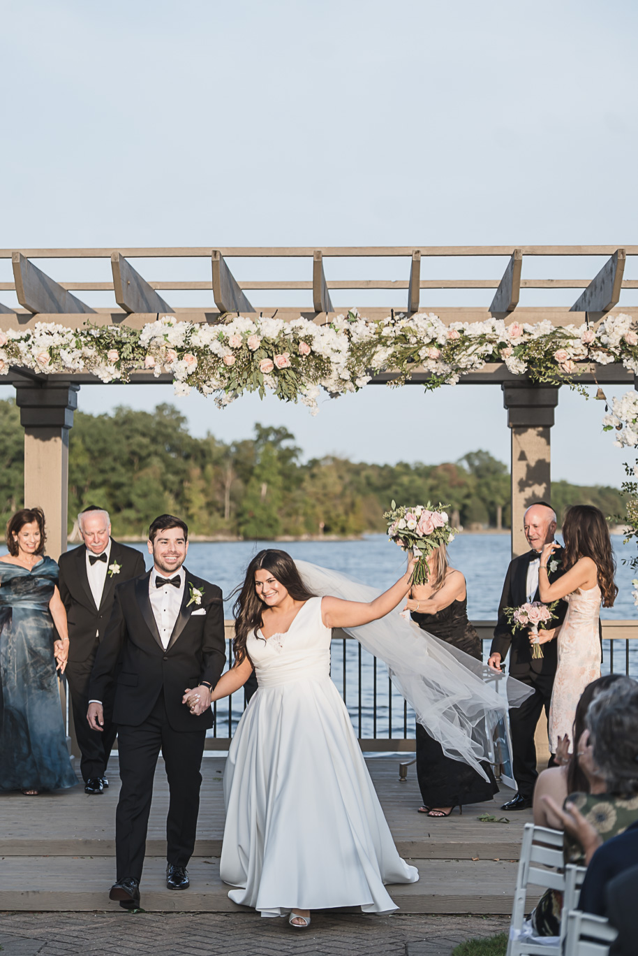 Newlyweds walk hand in hand down the aisle, smiling and celebrating beneath a floral chuppah at their Bay Pointe Golf Club Jewish wedding.