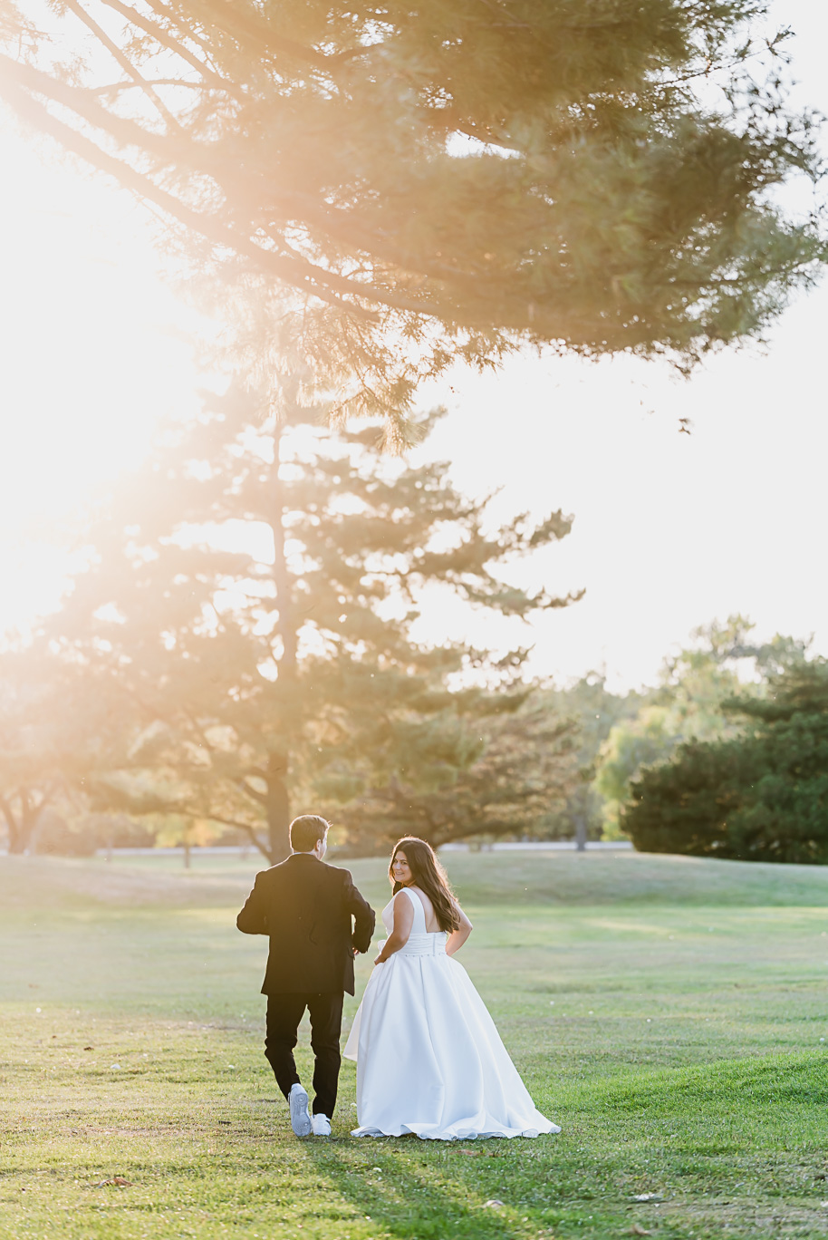 Bride in a classic white gown and groom in a black tuxedo walk hand in hand across Bay Pointe Golf Club at sunset, golden light glowing through the trees.