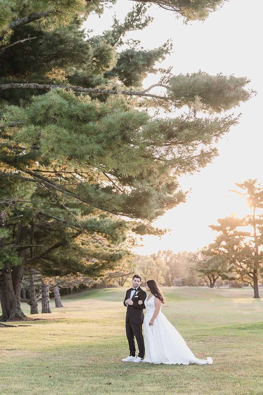 Bride in a classic white gown and groom in a black tuxedo walk hand in hand across Bay Pointe Golf Club at sunset, golden light glowing through the trees.