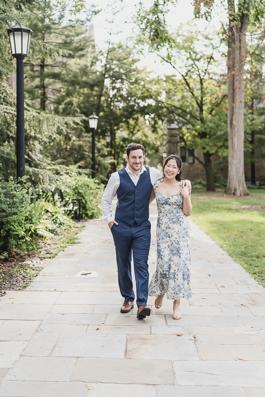 Linda and Alex walking arm-in-arm during their Michigan Law Quad engagement session, styled in a blue floral dress and navy vest suit, with romantic architecture and greenery as the backdrop.