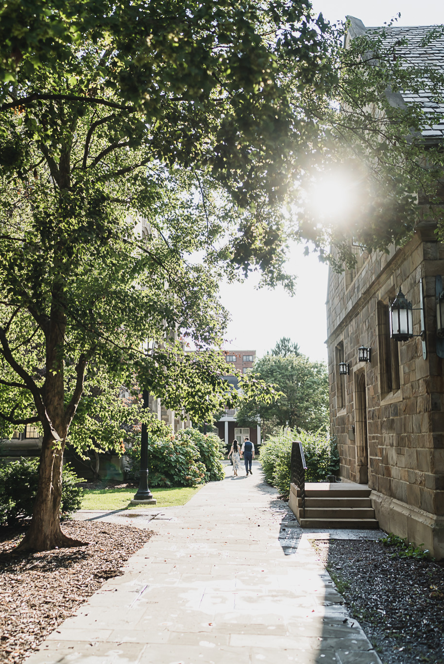 Sunlit engagement photo at the University of Michigan Law Quad with the couple walking hand-in-hand down a cobblestone path, captured with dreamy golden light by Ann Arbor wedding photographer Kari Dawson.