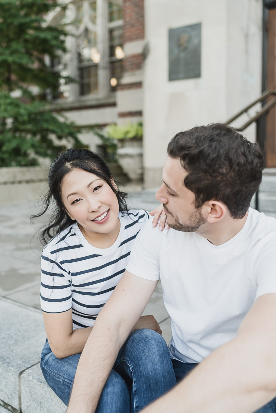 Engaged couple sitting together on the steps of the University of Michigan, bride-to-be in a striped top smiling lovingly at her fiancé during their Law Quad engagement session.