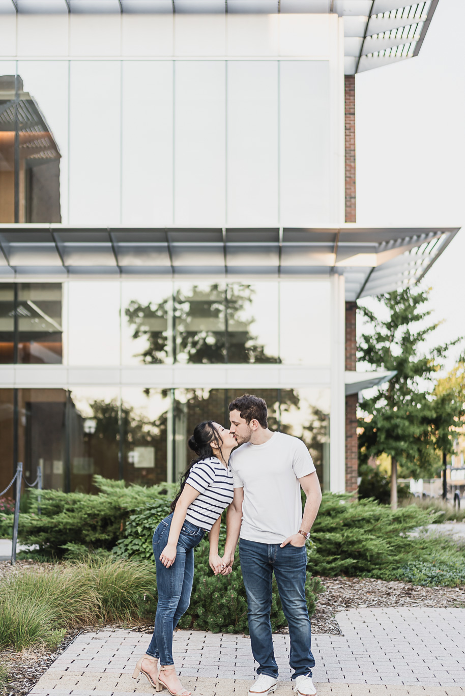 Romantic kiss framed by glass architecture at the University of Michigan, an elegant detail from their Michigan Law Quad engagement.