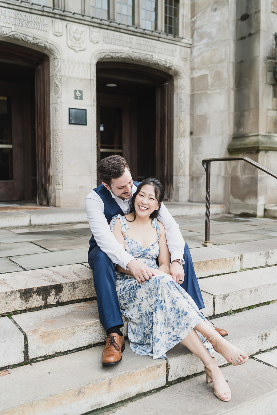 Romantic engagement photo at Michigan Law Quad featuring Linda in a floral dress and Alex in a navy vest, laughing together on the historic stone steps.