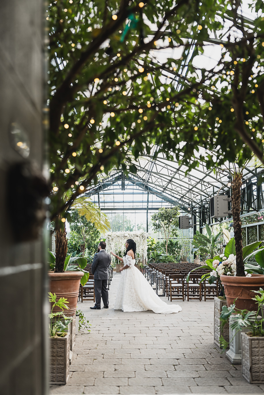 Bride and groom walk hand in hand through the glass greenhouse at Planterra Conservatory in Bloomfield Hills, Michigan, surrounded by lush greenery and soft autumn light during their elegant fall wedding.