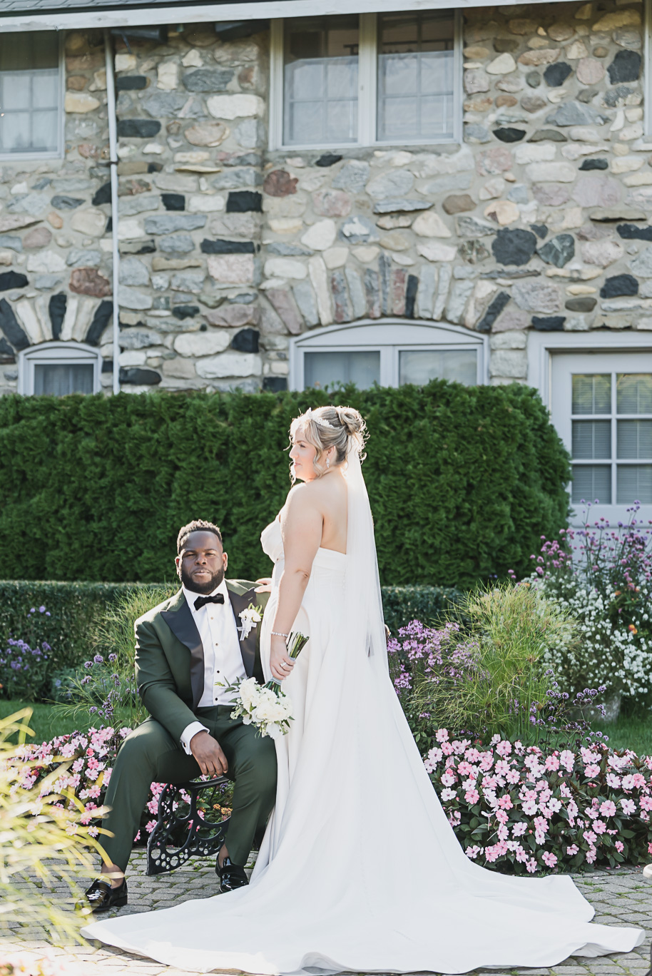“Editorial wedding portrait of an interracial couple at Castle Farms in Charlevoix, Northern Michigan. The bride in a strapless gown and cathedral veil stands beside her groom in an emerald green tuxedo, surrounded by classic stone architecture and soft garden florals. Captured by Kari Dawson + Co.
