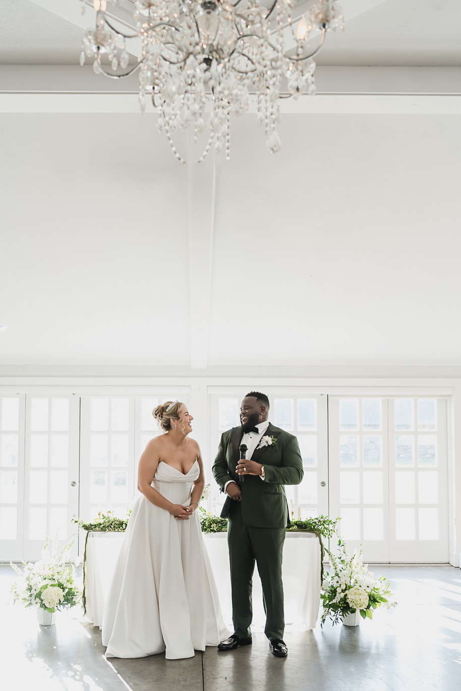 Joyful newlyweds sharing a heartfelt toast during their Castle Farms wedding reception in Charlevoix, Northern Michigan. Captured by Kari Dawson + Co, the bride in a strapless white gown and the groom in an emerald tuxedo laugh together beneath elegant chandeliers and soft natural light.