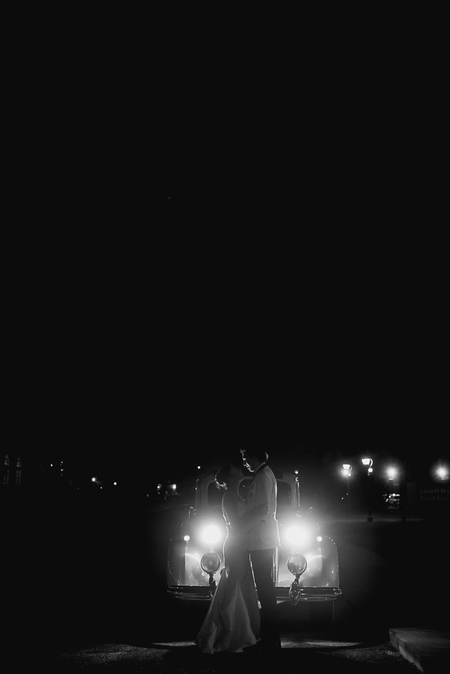 Newlyweds pose with their vintage ivory getaway car at night outside Lovett Hall, glowing with romantic lights.
