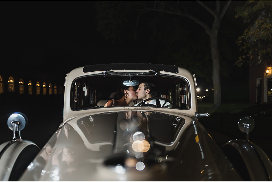 Newlyweds pose with their vintage ivory getaway car at night outside Lovett Hall, glowing with romantic lights.
