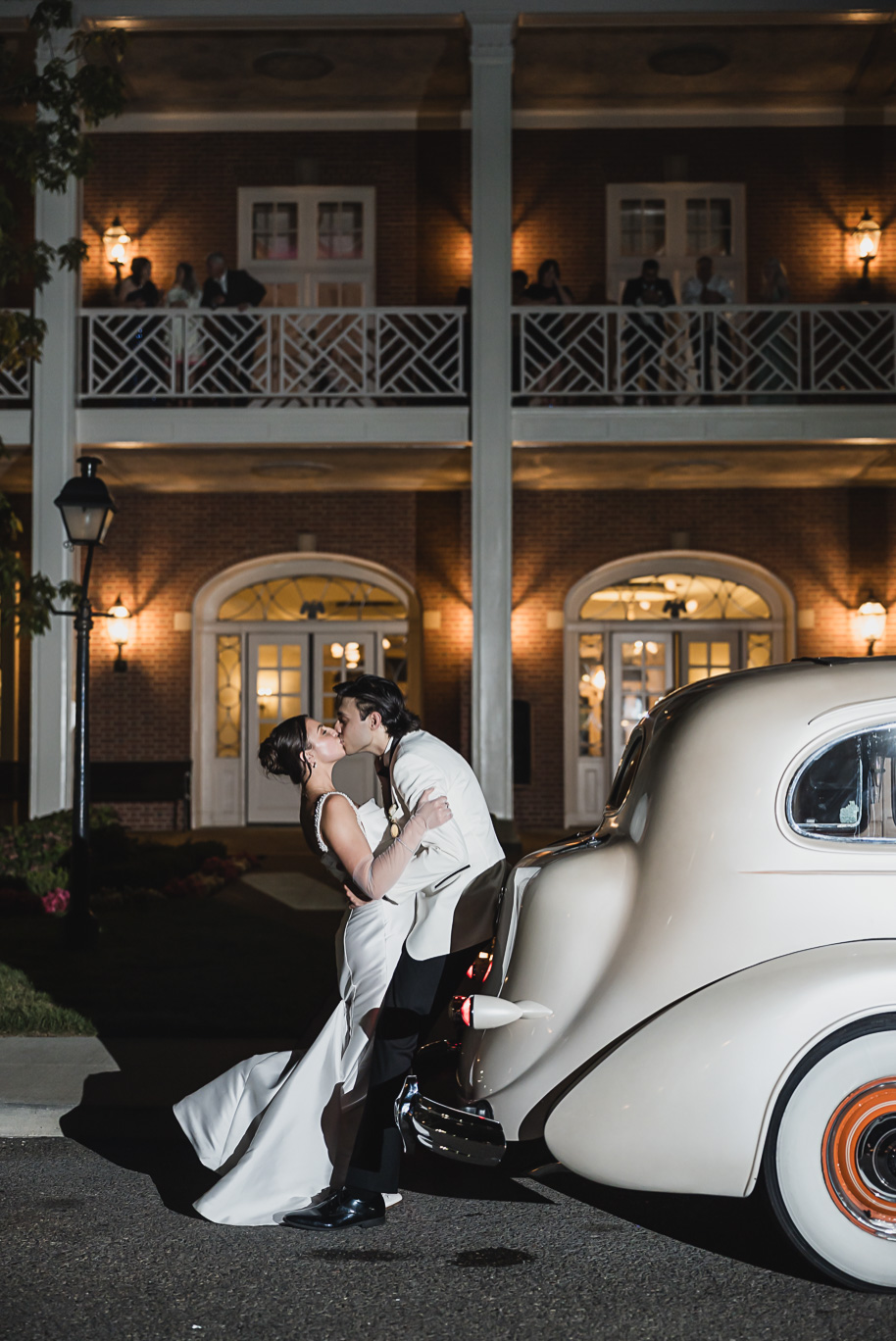 Newlyweds pose with their vintage ivory getaway car at night outside Lovett Hall, glowing with romantic lights.

