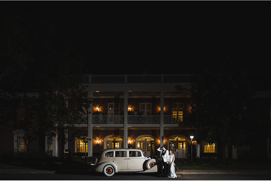 Newlyweds pose with their vintage ivory getaway car at night outside Lovett Hall, glowing with romantic lights.
