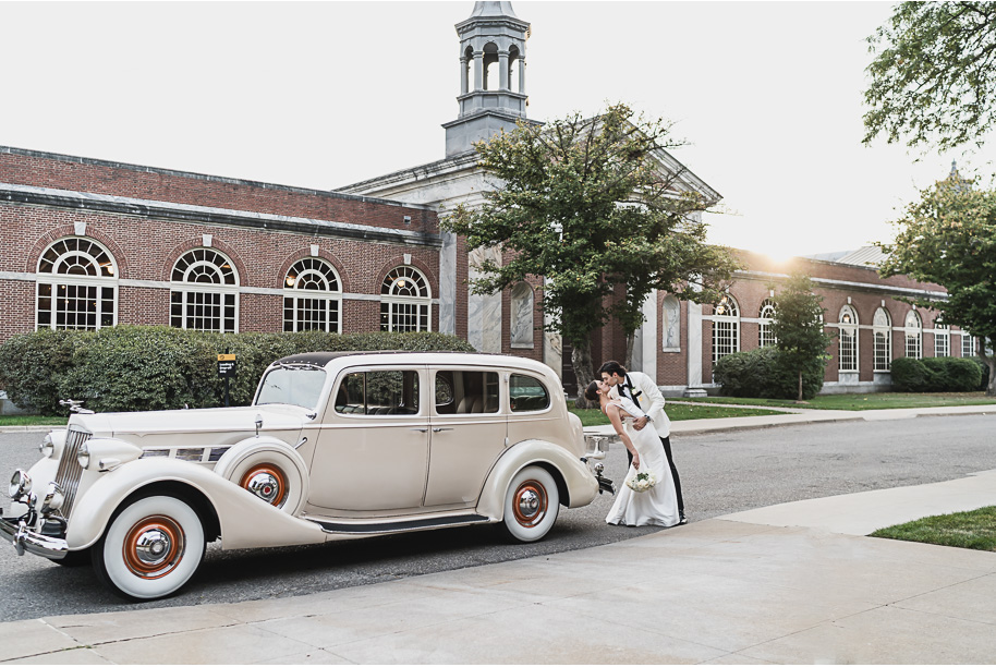 Newlyweds pose with their vintage ivory getaway car at night outside Lovett Hall, glowing with romantic lights.
