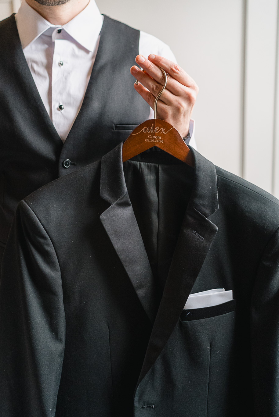 A custom black tuxedo for a groom on his wedding day along with a custom wood hanger engraved with Alex, groom, and wedding date.